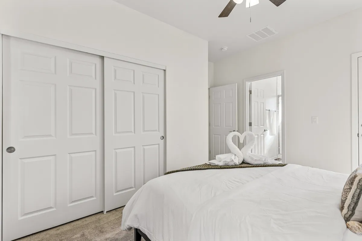 A neatly made queen bed with towels arranged in a heart shape sits beside closet doors and a mirrored wall. The gentle lighting and thoughtful gestures foster a romantic, serene mood.