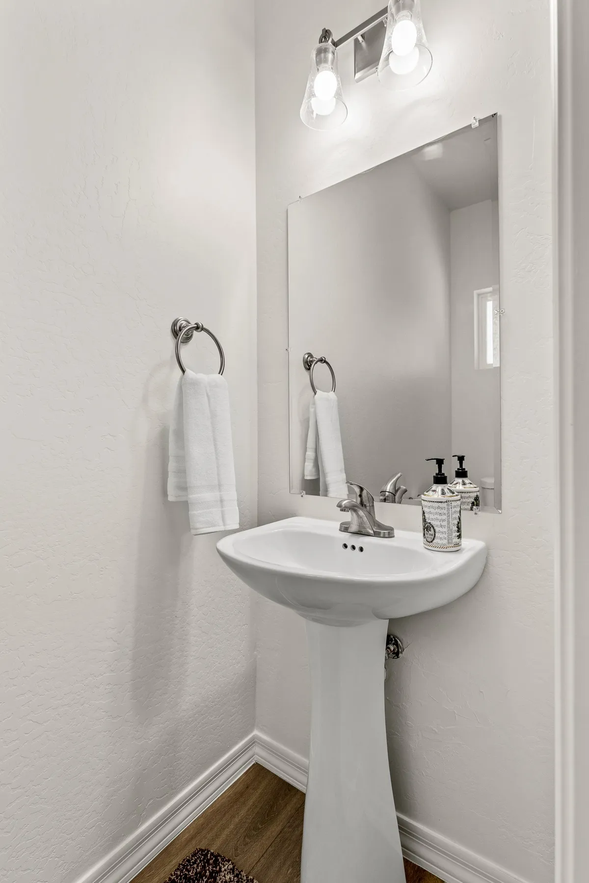 A well-lit half bathroom featuring a pedestal sink, mirror, towel rack, and modern light fixtures. The space exudes cleanliness and hotel-inspired serenity.