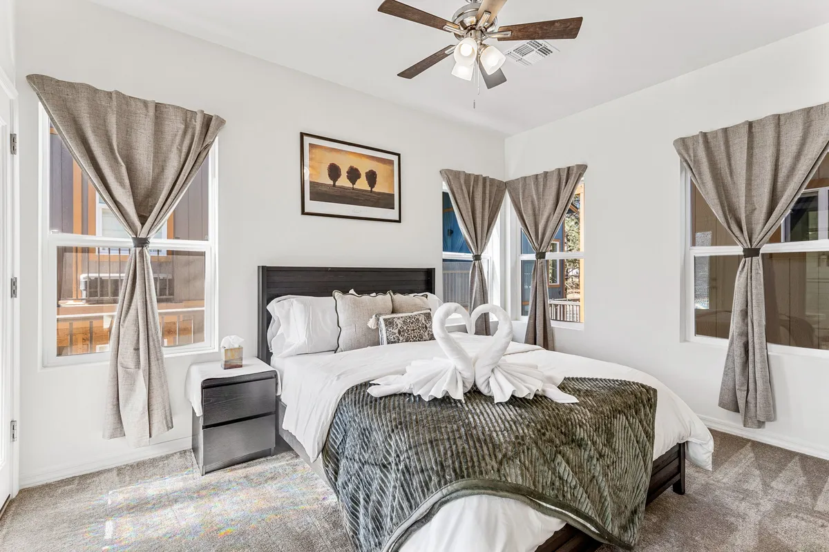 The image features a beautifully made queen bed with decorative towels shaped like swans, soft ambient lighting, tasteful wall art, and elegant window treatments letting in gentle sunlight. The overall mood is romantic and serene, inviting relaxation and comfort.