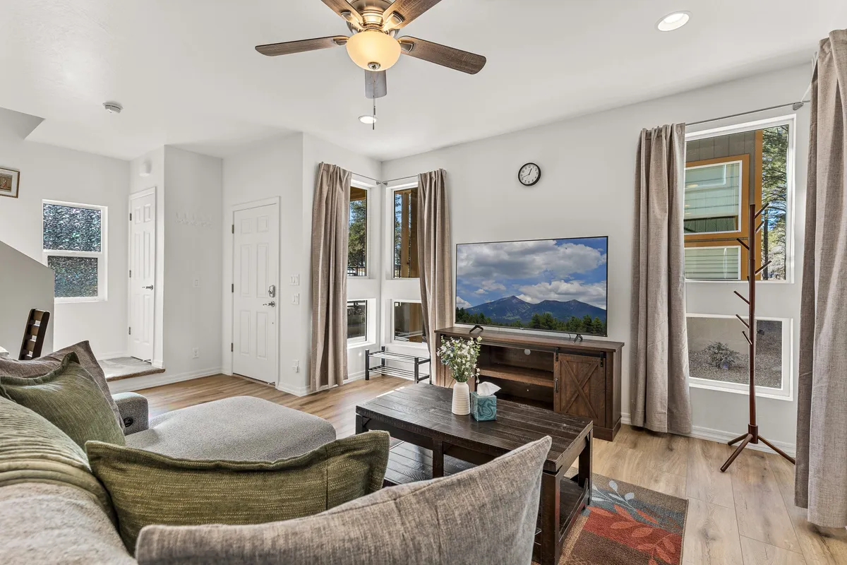 A cozy living space with a comfortable sofa, coffee table, TV, ceiling fan, and large windows. The windows provide glimpses of the outdoors, suggesting a seamless flow between interior comfort and nature beyond. The setup hints at relaxing with a morning coffee or watching a sunset through the windows, echoing the atmosphere described for the balcony.
