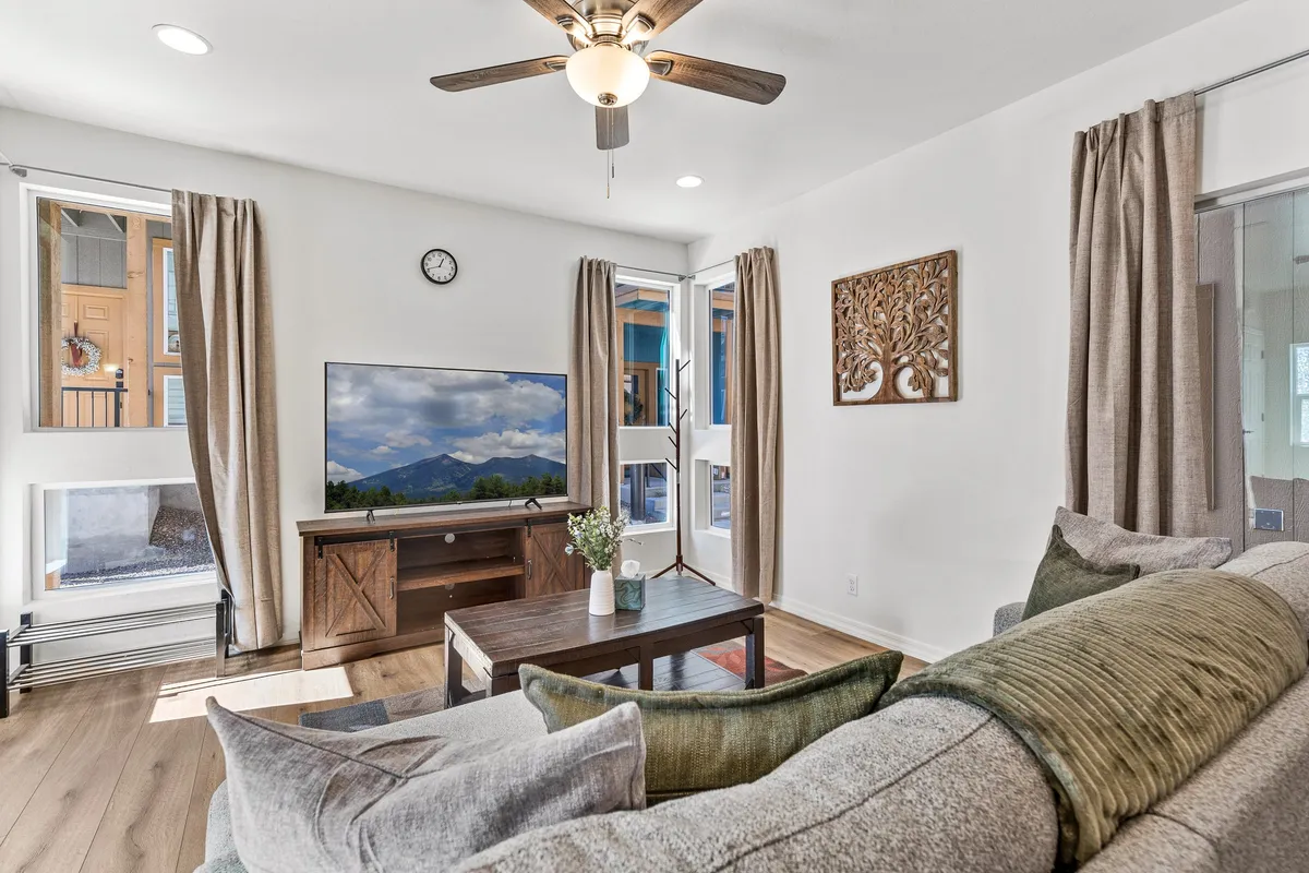 A cozy and comfortable living room featuring a plush sofa adorned with decorative pillows, a coffee table with a vase of flowers, a television displaying a scenic view, and ample natural light streaming in through a large window. The space feels inviting and makes it easy to unwind after a day outdoors.