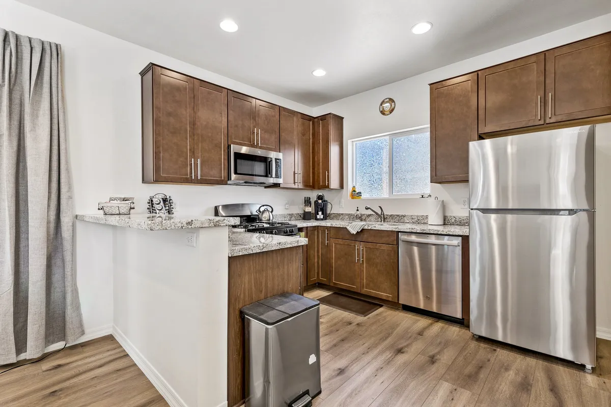 A warm, functional kitchen scene with granite countertops, stainless steel appliances, wooden cabinets, and a window letting in natural light. The space is clearly set up for cooking, entertaining, and sharing meals together.