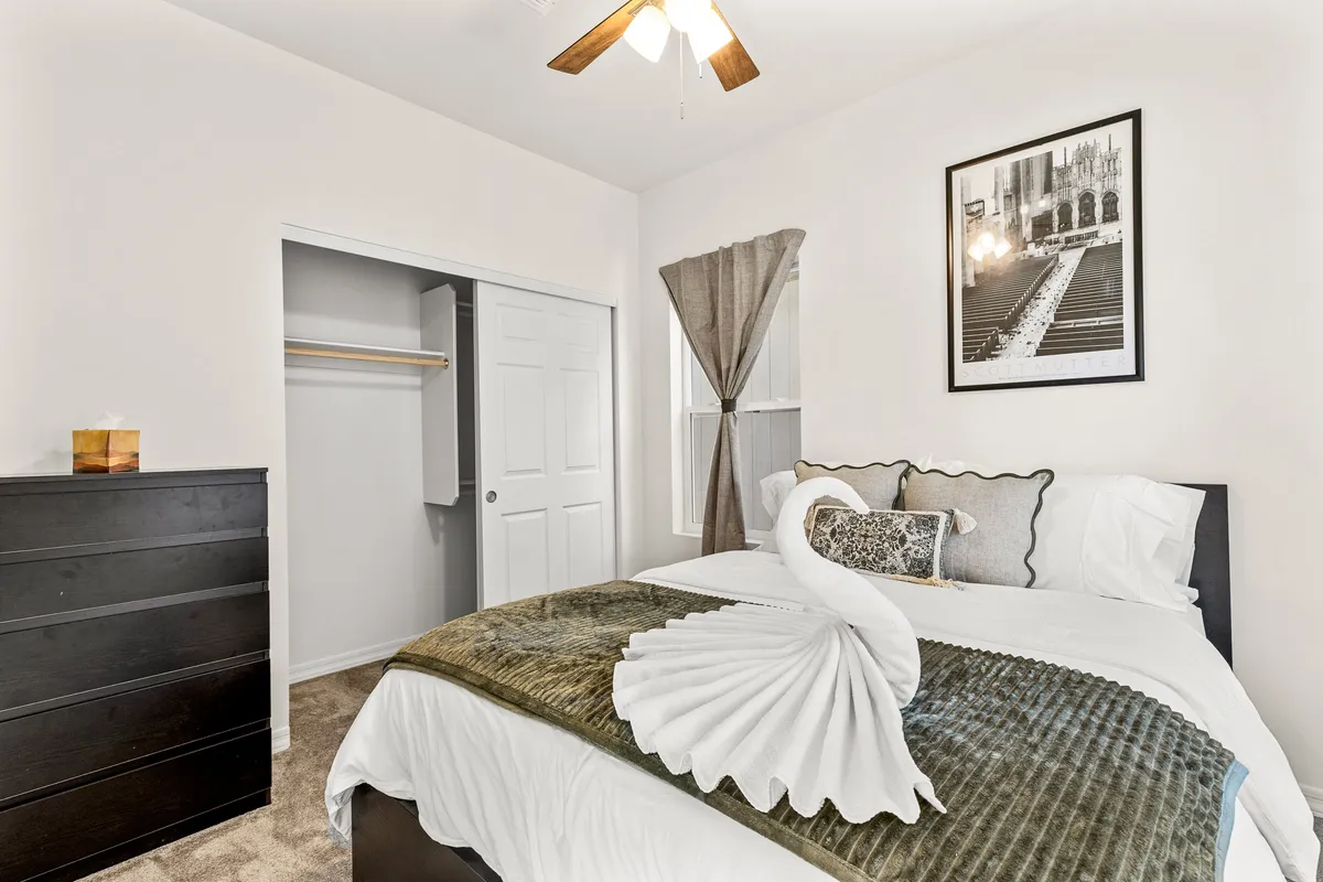 A serene bedroom featuring a plush queen bed adorned with a decorative swan towel, a dresser, and a framed black-and-white photograph. Soft natural light filters in through a window, contributing to the calming, restful atmosphere.